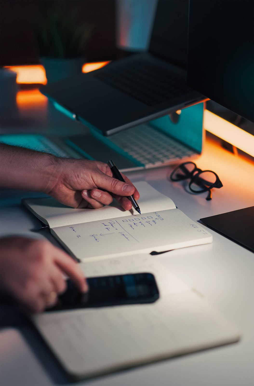 Hands writing a schedule in a notebook beside a laptop on a dark desk
