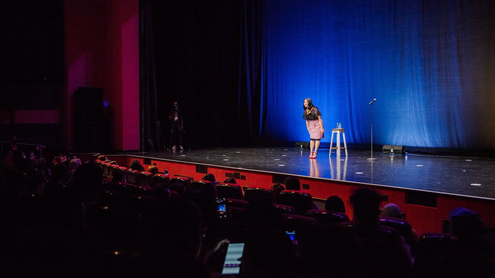 Stand-up comedian alone on a large theatre stage under blue light, seen from the audience